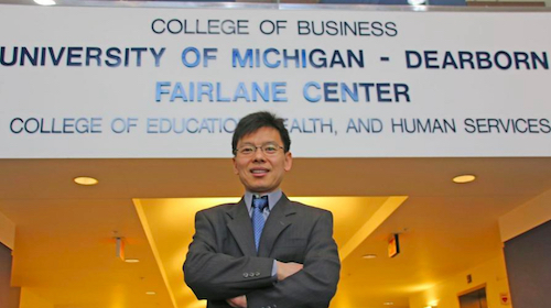 Jun He standing in front of wall that reads: College of Business, University of Michigan-Dearborn, Fairlane Center, College of Education, Health, and Human Services