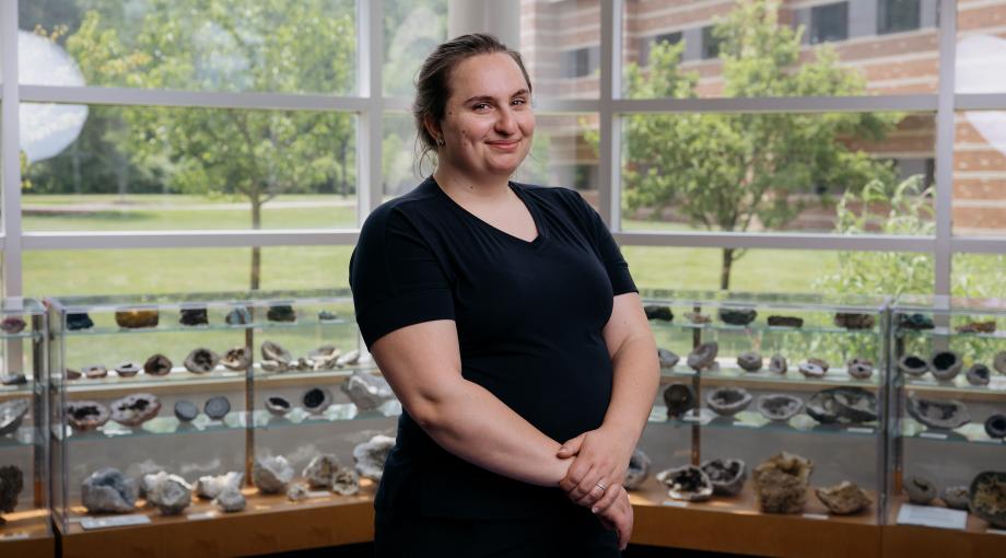UM-Dearborn alum Kristen Dage stands for a formal portrait in the Natural Sciences Building atrium at UM-Dearborn, with windows and artifact display cases in the background.