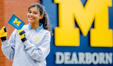 A smiling students holds up a small Block M flag in front of a large UM-Dearborn Block M logo.
