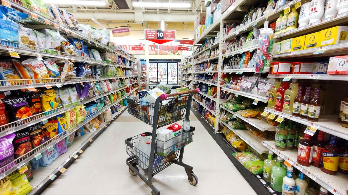 A shopping cart full of groceries sits in the middle of the snack food aisle at a brightly light grocery store.