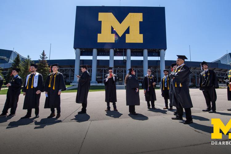Graduates in academic gear stand outside Michigan Stadium, the Big House