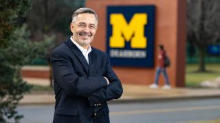 Chancellor Domenico Grass stands for a portrait with his arms crossed in front of a Block M UM-Dearborn logo on the UM-Dearborn campus.