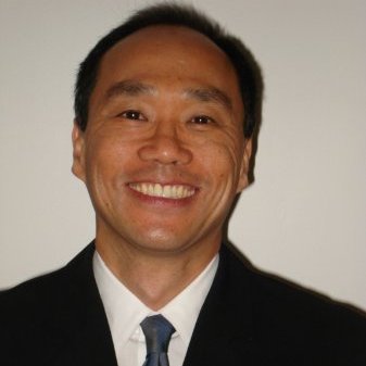 A photo of David Hay. He has short black hair and dark brown eyes. He is smiling widely for the camera. He is wearing a black suit with a blue tie and white shirt underneath. 