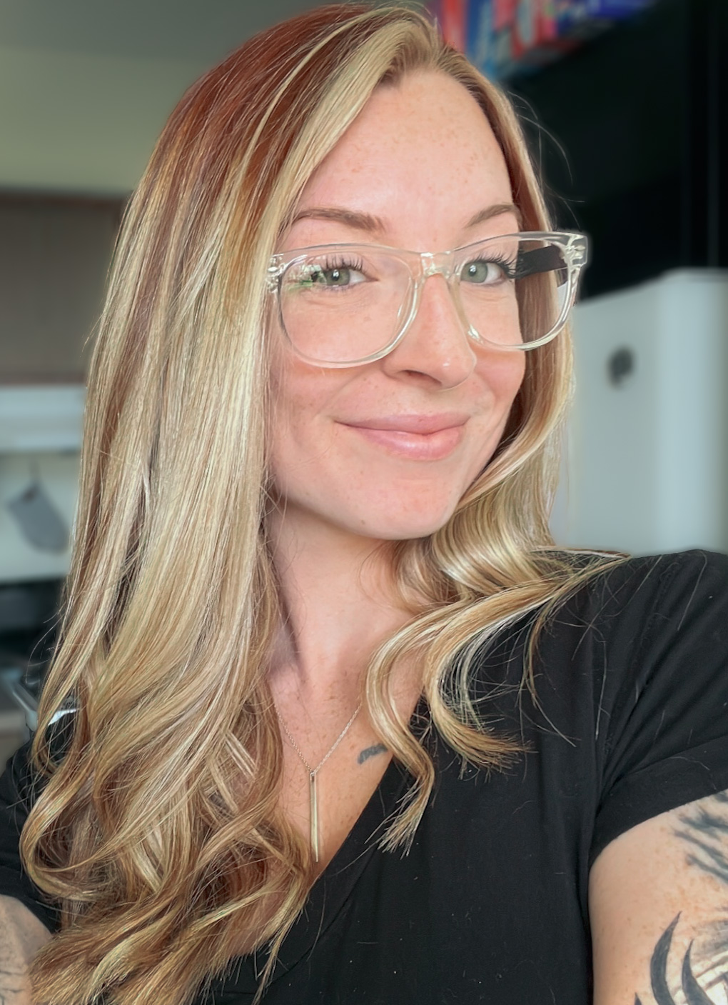 A photo of Becca Jarvis. She has long blonde hair and green eyes and is wearing clear framed glasses and a black tee shirt. She has a tattoo showing on her arm.