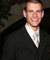 A photo of Bradley Brennan in a black pinstripe suit, white button up, and silver-gray tie.