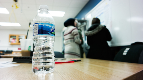 Bottled water sitting on a desk with students in the background writing on a whiteboard.