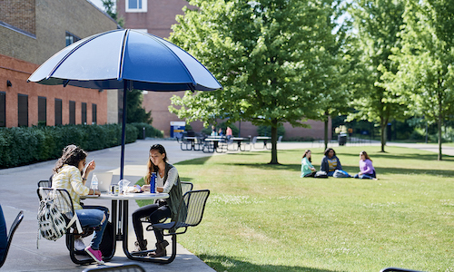 Two students sitting an outdoor table under an umbrella on campus.