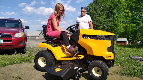  A young woman in a dress and work boots sits on a riding lawnmower, flanked by an older man who is explaining how to use it. 