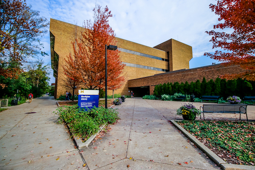  A fall exterior view of the Mardigian Library on the UM-Dearborn campus, with a blazing red maple tree in the foreground and leaves blowing over the sidewalks. 