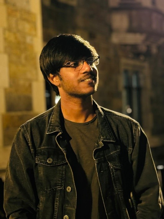 Gokul is looking to his left wearing a black denim jacket, glasses, and is smiling