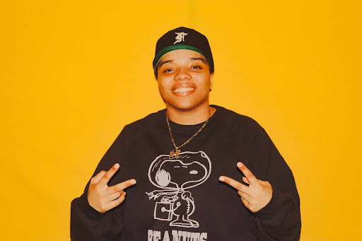 sydney is in front of a yellow background in a black sweater and black cap, smiling and holding up two peace signs