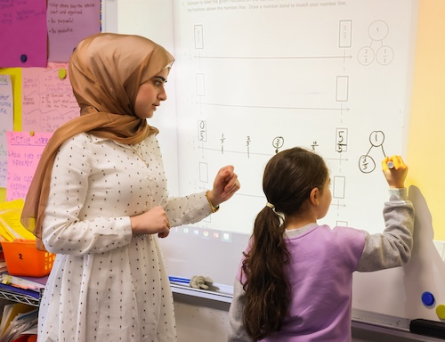 Student teacher watching over young student at white board