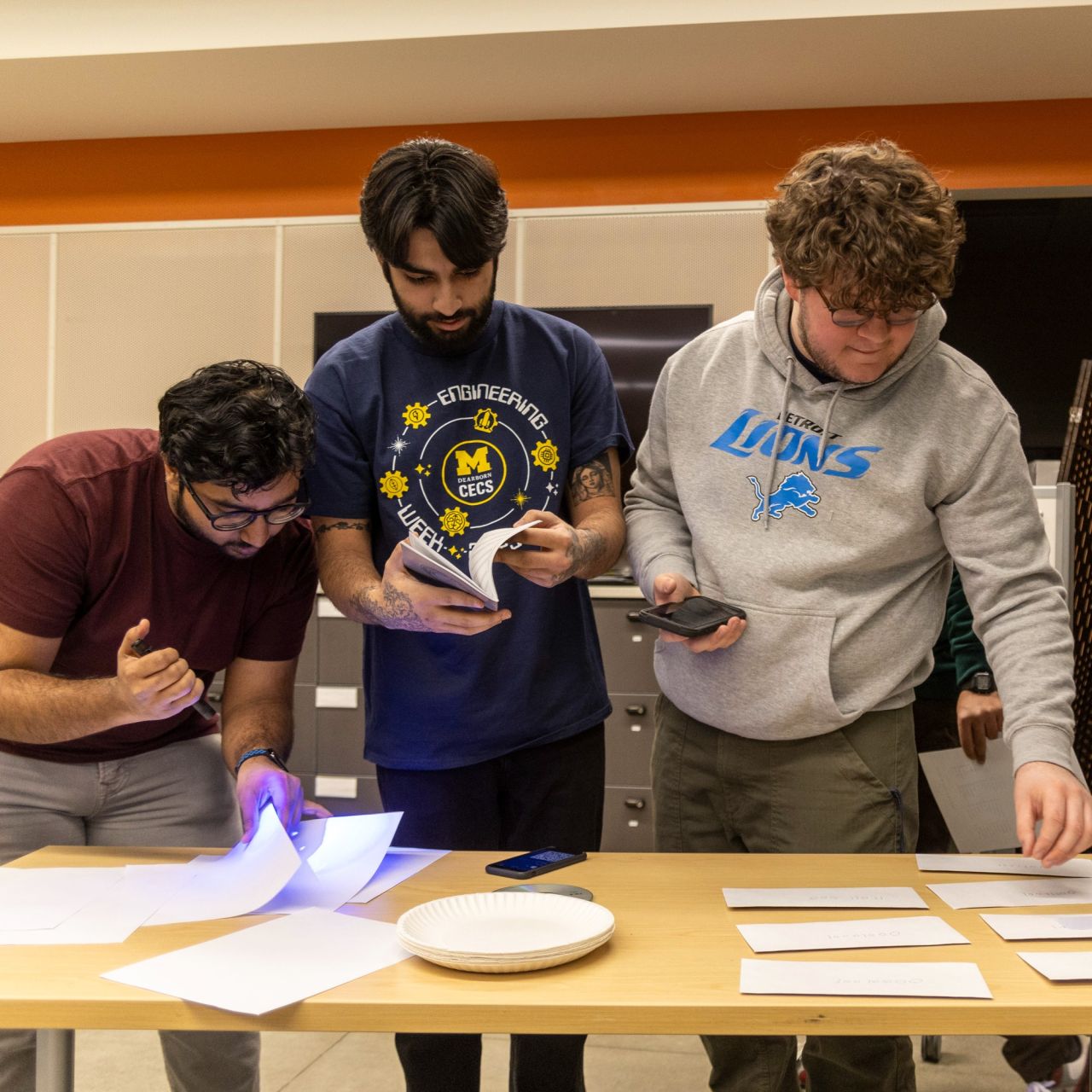 Three individuals are examining and sorting documents on a table in a brightly lit room. One person is wearing a "Michigan Engineering" T-shirt, and another sports a "Lions" hoodie. All are focused on their tasks, handling papers and using a mobile device.