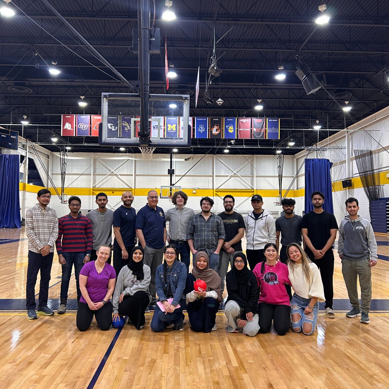 Group of people posing in a basketball gymnasium with visible team banners hanging in the background.