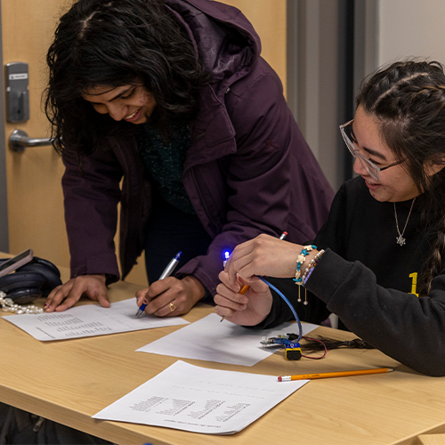 Two students are engaged in studying and writing notes at a desk in a classroom setting.