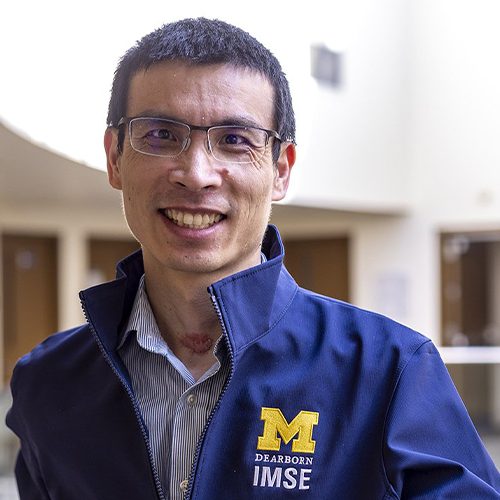 Smiling individual wearing a University of Michigan-Dearborn IMSE jacket standing in a brightly lit atrium.