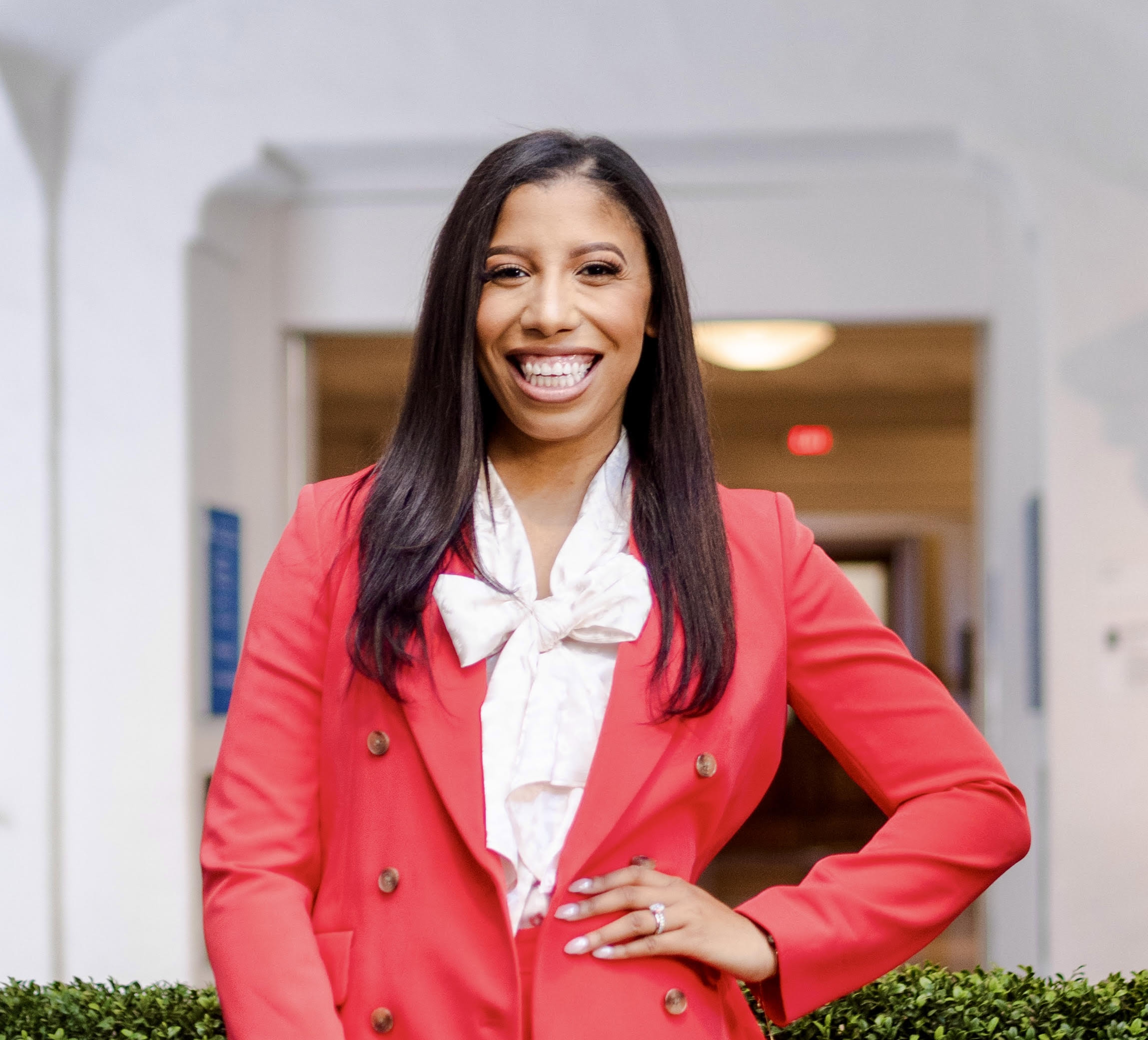A photo of Kimberly wearing a red suitcoat with a white frilled bow underneath standing in front of a white building
