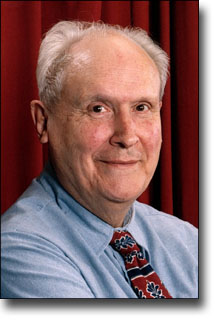 Portrait of person, smiling, wearing a blue shirt and a tie with a celestial pattern, set against a red curtain background.