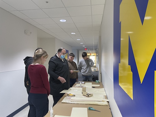 A group of students gathered around a table in a hallway facing a wall with UM-Dearborns M-logo on it, with Gerald Ottenbreit showing how to make boxes for fragile books. 