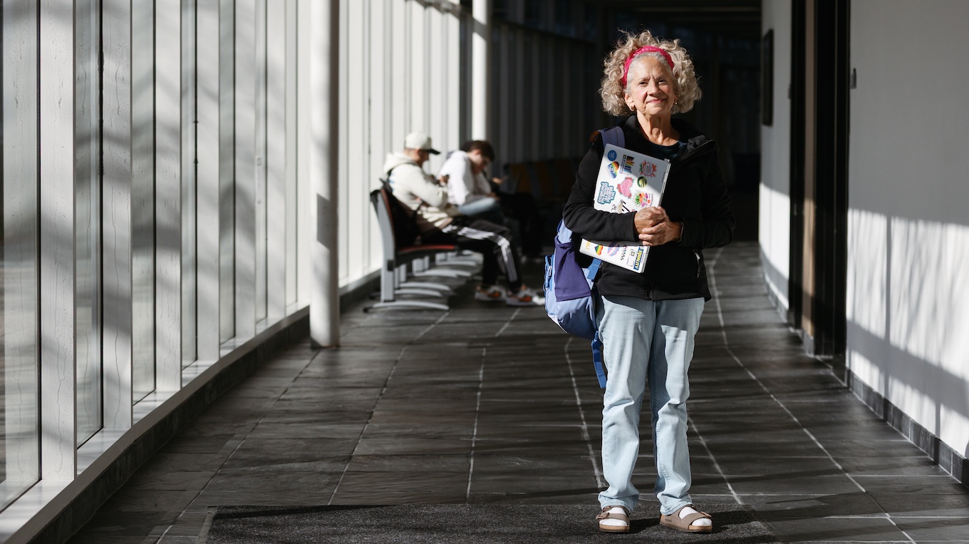 An older woman stands in a hallway lined with windows while holding a laptop computer.