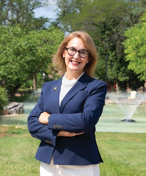 Chancellor Scarlatta wearing glasses, a navy blazer, and a pearl necklace stands confidently with arms crossed in front of a fountain surrounded by lush green trees and grass on a sunny day.