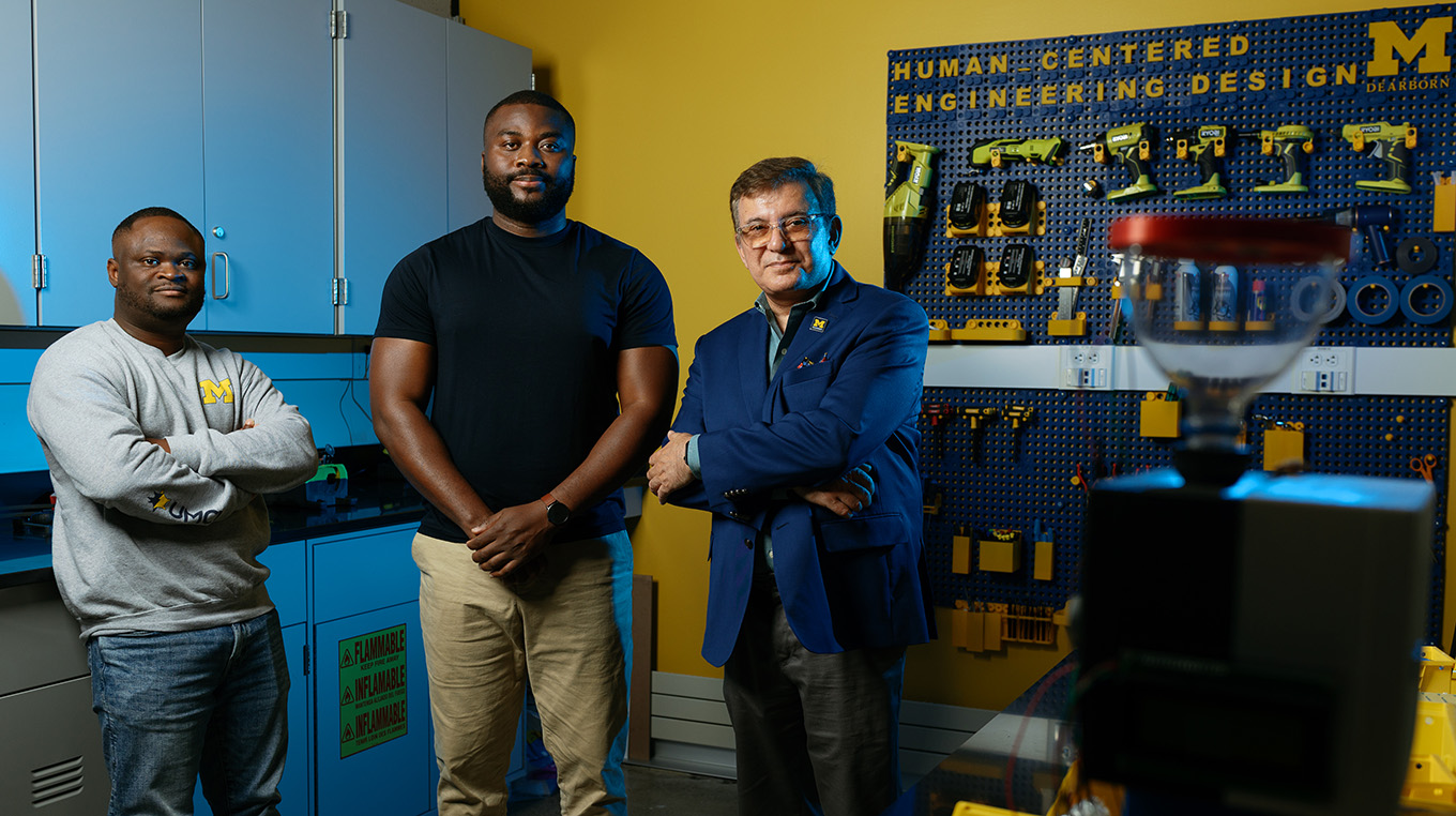 Two graduate students stand next to a professor in a university manufacturing lab, decked out in Maize and Blue.