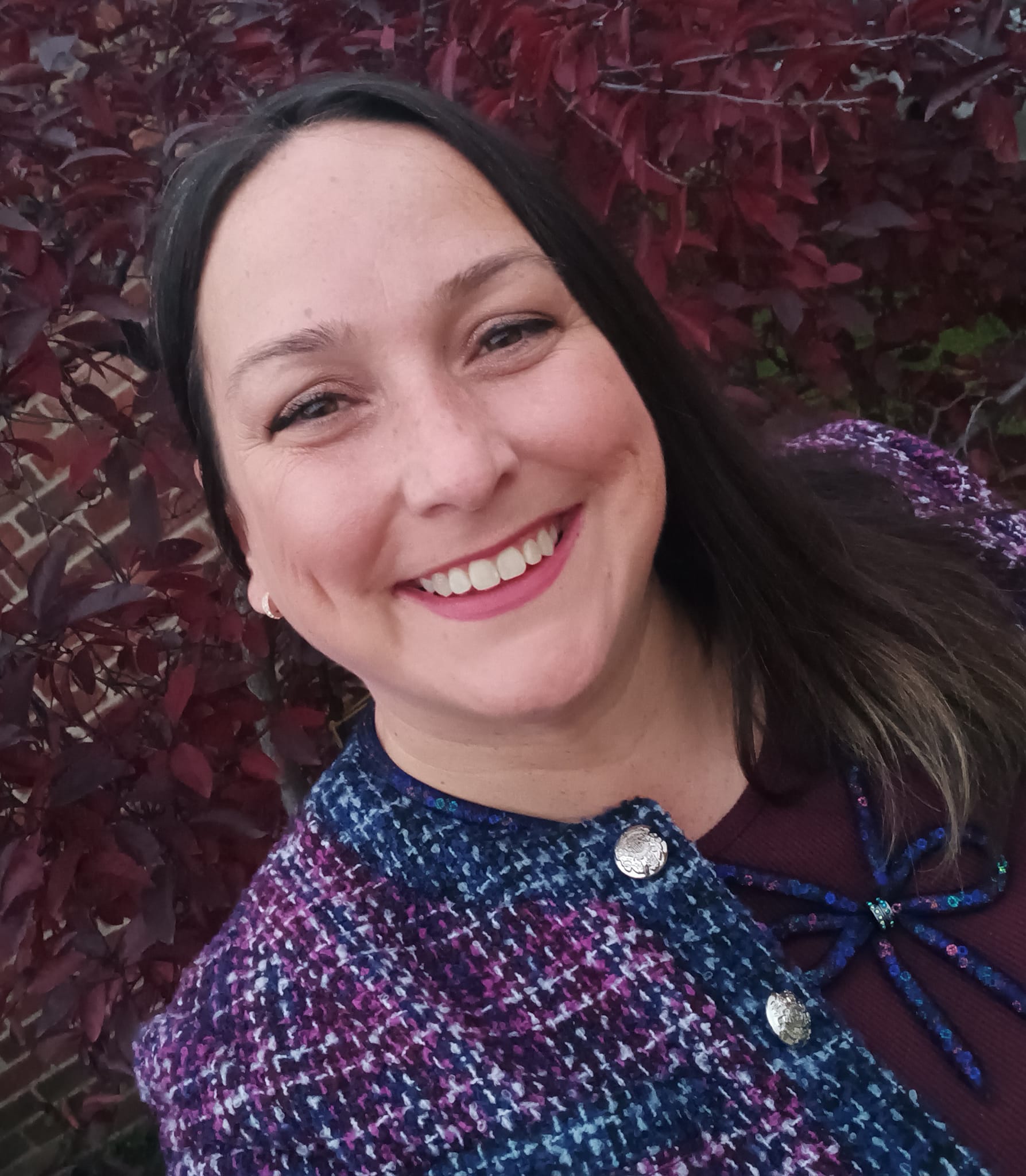 Person wearing a purple and blue textured jacket with decorative buttons.Standing outdoors near a bush with red leaves. Brick wall visible in the background.