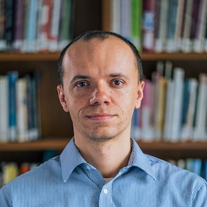 A photo of a man in a  blue shirt looking forward. He is in front of a book shelf