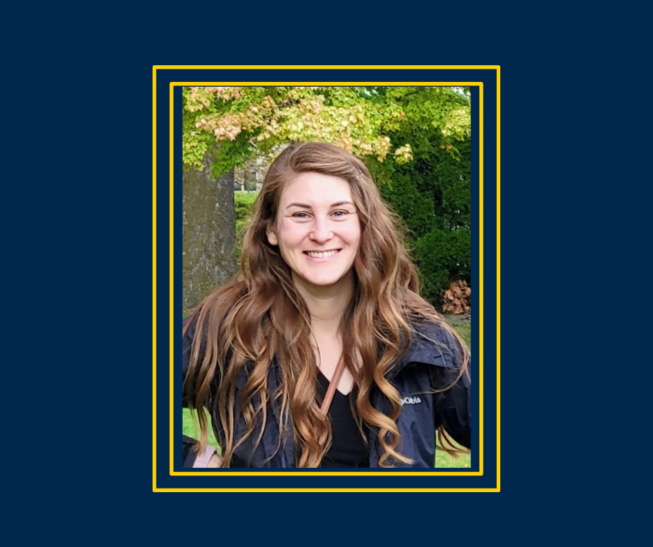 Smiling woman with long wavy hair standing outdoors in front of green trees, framed by a maize and blue border.