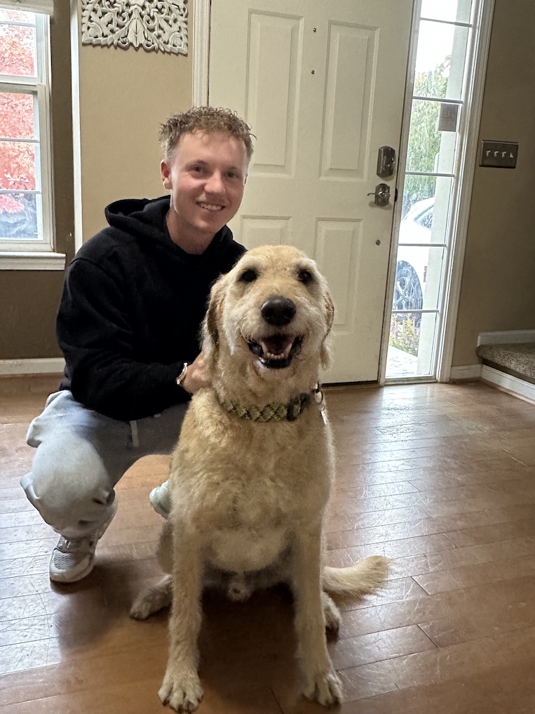 A smiling man kneels next to his beige-colored smiling dog.