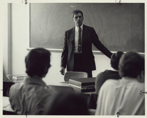 A man dressed in a suit stands in front of a blackboard to teach students in a black and white photo.