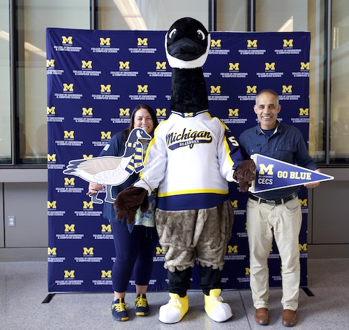 Two people standing with the University of Michigan mascot in a lobby, all wearing Michigan sports attire. One person holds a 'Go Blue' sign.