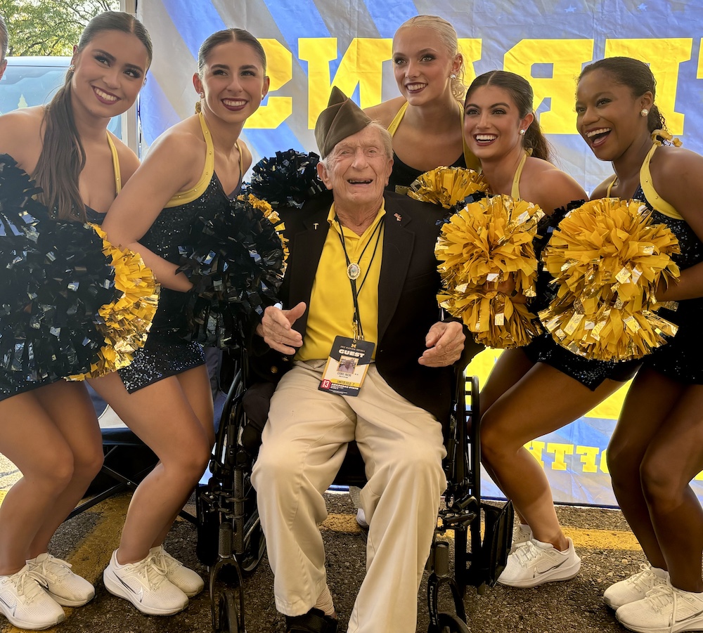 Female cheerleaders in maize and blue surround an older gentleman in a military uniform who is seated in a wheelchair
