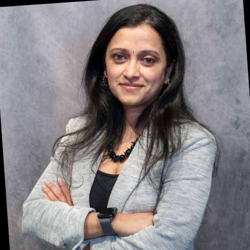Portrait of a smiling individual with crossed arms, wearing a gray blazer and black necklace, standing against a textured gray background.