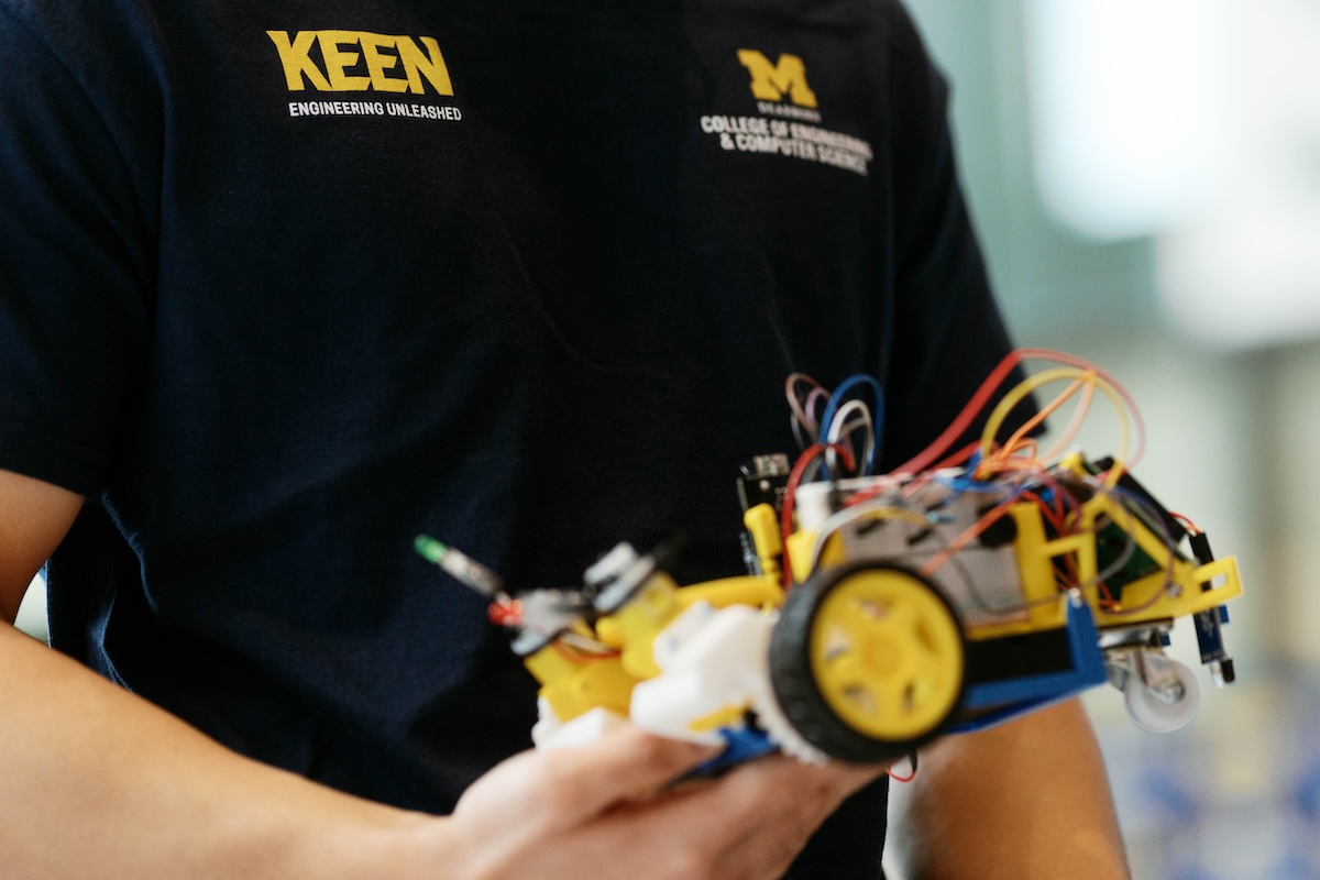 Person holding a small robotic vehicle with wires and sensors, wearing a KEEN Engineering Unleashed and University of Michigan t-shirt.Person holding a small robotic vehicle with wires and sensors, wearing a KEEN Engineering Unleashed and University of Michigan t-shirt.