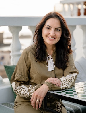 A smiling woman wearing beige is seated outdoors with a white fence behind her.