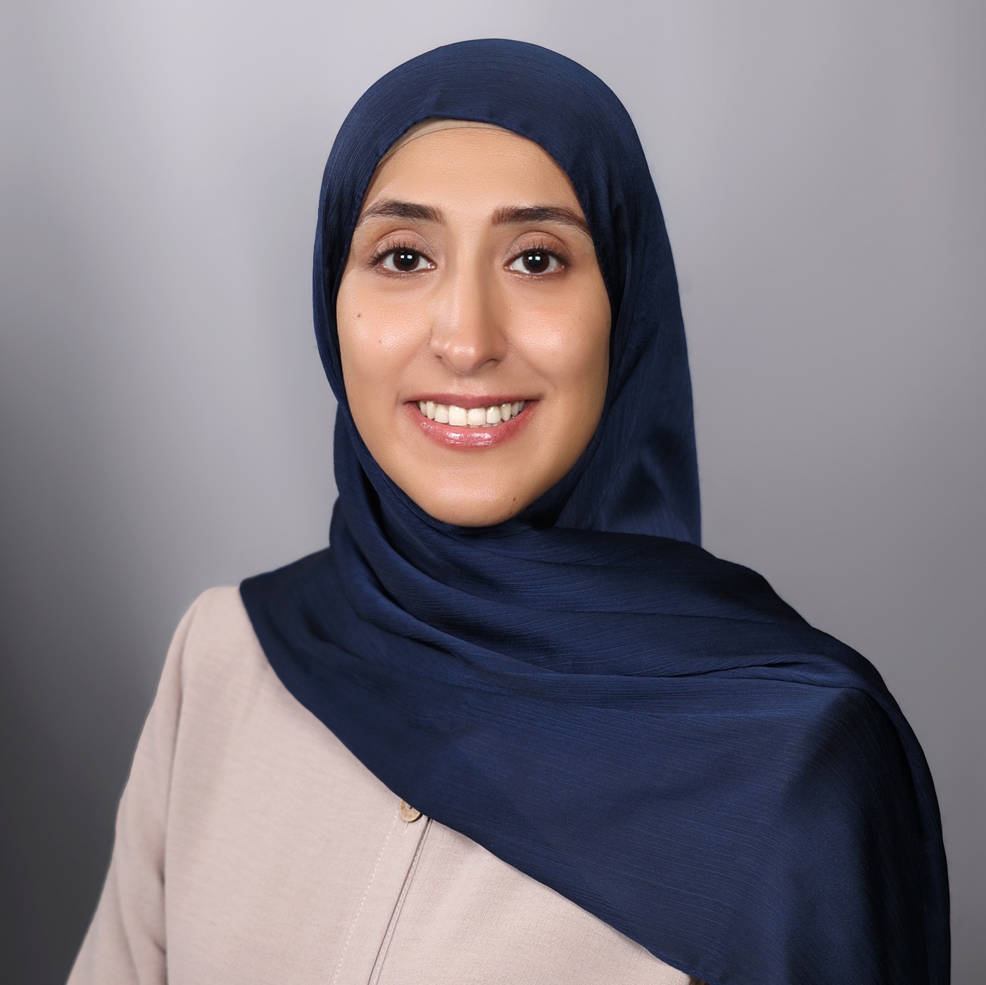 A smiling woman wearing a blue hijab and beige dress poses for a portrait