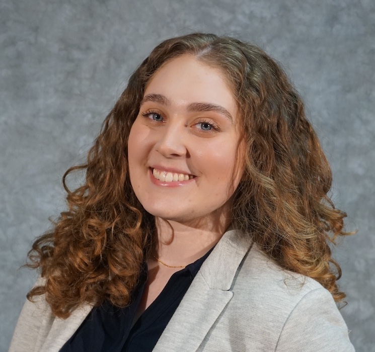 A photo of a brown curly-haired woman wearing a gray suit jacket and a black shirt