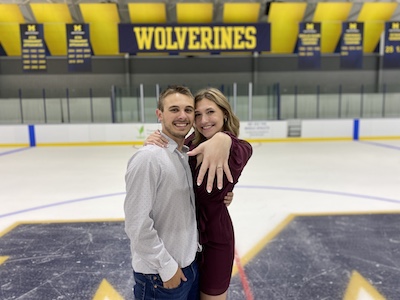 A man and woman stand on the ice with a maize and blue sign that reads "Wolverines" behind them.