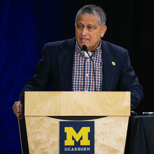 A man wearing a red, white and blue checkered button-up shirt and navy blue suit jacket speaks at a podium.