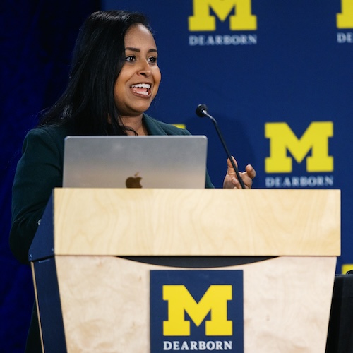 A woman wearing a teal blue suit jacket and a black shirt speaks to an audience from a podium