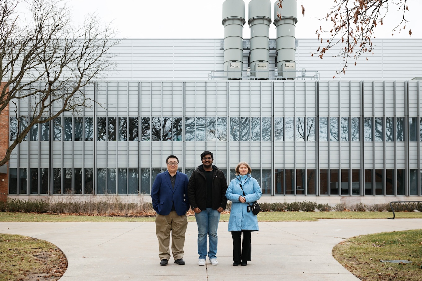 Three people — two males and a female — stand in front of a university building. The building is silver and has three smokestacks.