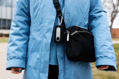 A close-up photo of a rectangular black air quality sensor that a woman is wearing on her black purse. The woman is wearing a bright blue coat.