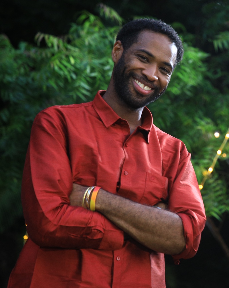 A photo of Jerrard Wheeler in a red shirt in front of a dark green leafy background with string lights.