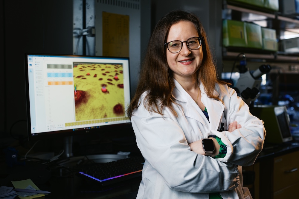 A photo of a female professor in a while lab coat. She is standing in front of a laptop monitor that shows her cancer research.
