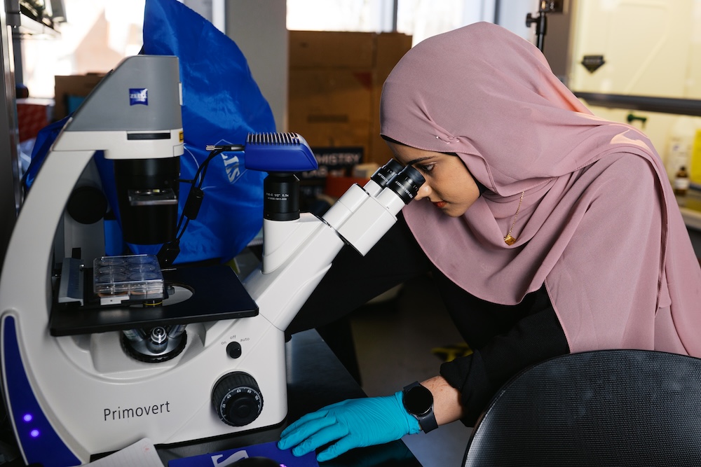 A female student researcher looks at melanoma tumor growth through a white microscope.