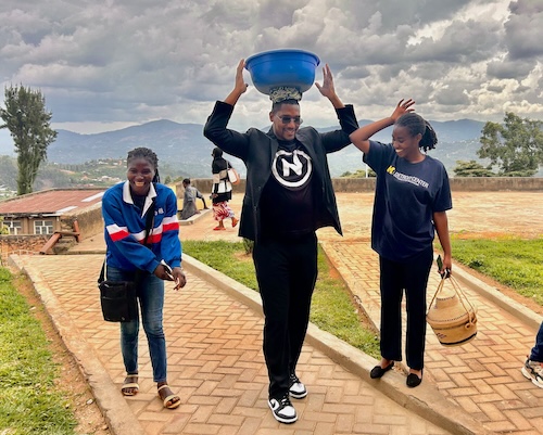 Three people laugh while they walk up a dirt road. The one in the center has a large blue basket on his head. There are avocados in the basket.
