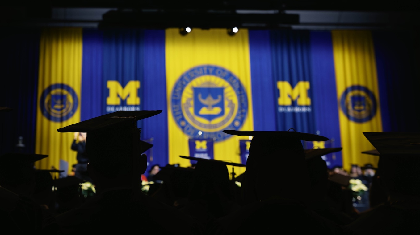 Silhouettes of graduates face a stage draped in maize-and-blue banners with University of Michigan-Dearborn logos and the university seal at center.