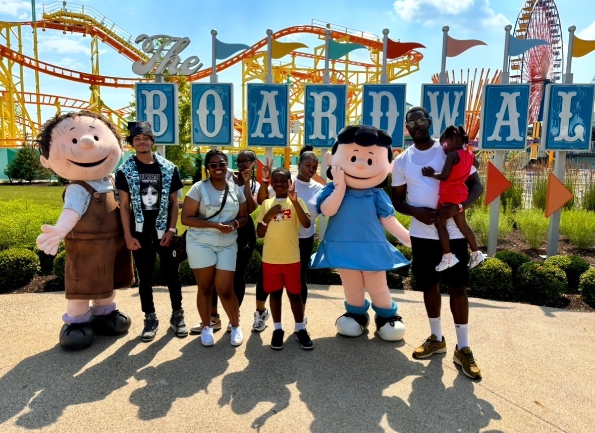 A group of two adults and five children visited an amusement park. They are standing with Peanuts cartoon characters with a roller coaster in the background