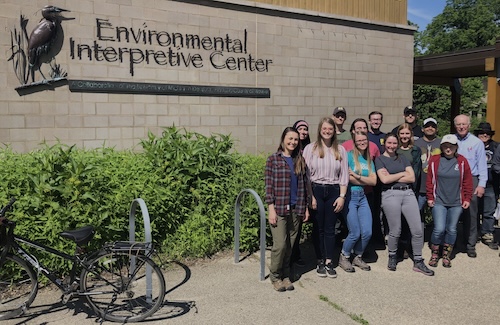 A group of students and a professor stand to the right of a light grey-ish beige brick building. The building was a sign that reads, "Environmental Interpretative Center.